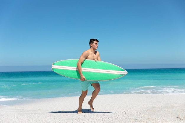 Vista lateral de cerca de un hombre caucásico caminando por la playa con el cielo azul y el mar en el fondo, llevando una tabla de surf