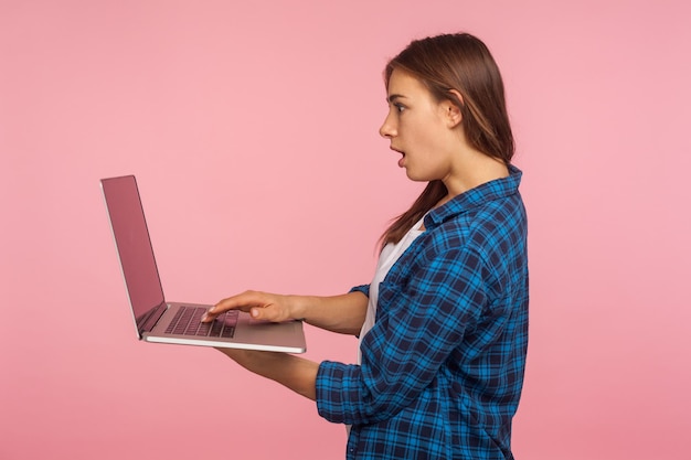 Vista lateral de una chica asombrada con un pantalones casual a cuadros leyendo correos electrónicos de una laptop con expresión asombrada, usando una computadora para educación o trabajo en línea, navegando por Internet. tiro de estudio interior, aislado