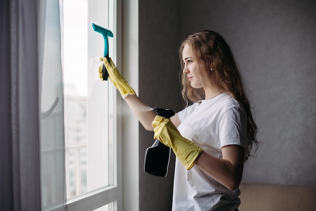 Vista lateral de una chica rizada con camisa blanca y guantes protectores amarillos con una ventana especial de limpieza de cepillos Empleada de la empresa de limpieza con uniforme trabajando sola