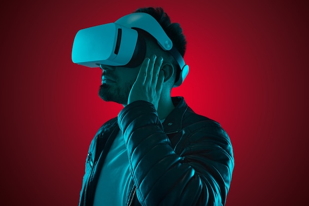 Vista lateral del chico barbudo contemporáneo con chaqueta de cuero y gafas de realidad virtual ajustando los auriculares TWS mientras ve videos en estudio con luces de neón