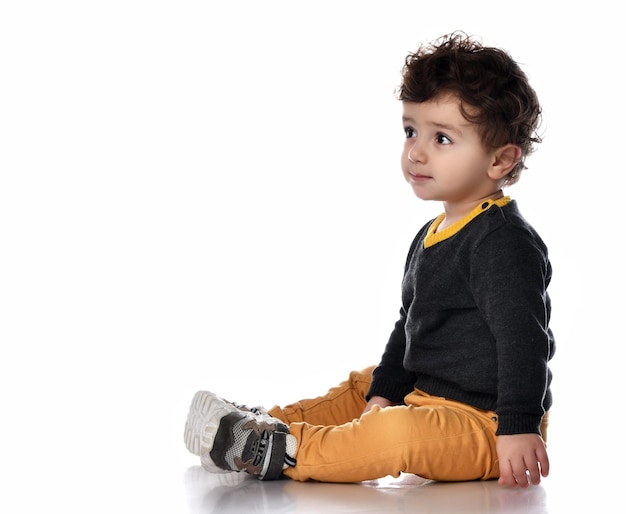 Vista lateral de un chico guapo y elegante sentado en un fondo blanco y mirando hacia un lado con interés Un niño con pantalones de suéter y zapatillas se divierte en el estudio