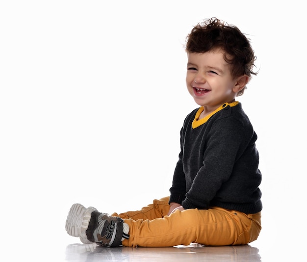 Vista lateral de un chico guapo y elegante sentado sobre un fondo blanco y riéndose alegremente Un niño con pantalones de suéter y zapatillas de deporte se está divirtiendo en el estudio Concepto de emociones infantiles