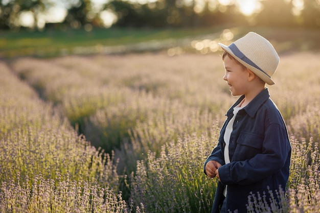 Vista lateral de un chico lindo con sombrero caminando en un campo de lavanda al atardecer