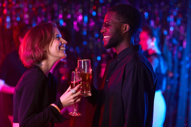 Vista lateral de la cintura hacia arriba en una pareja joven sonriente disfrutando de bebidas en la fiesta con luz de neón