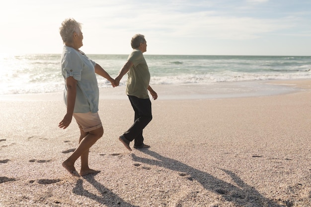 Vista lateral completa de una pareja multirracial de ancianos cogidos de la mano mientras camina por la orilla de la playa