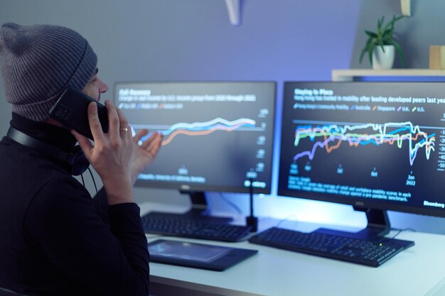 Vista lateral del corredor de bolsa hipster hombre hablando por teléfono móvil con gráficos de análisis en múltiples pantallas de computadora en conversaciones de fondo con su socio comercial