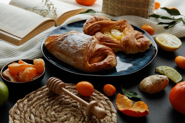 Vista lateral de croissants en plato con rodajas de mandarina y libro abierto con cesta de kumquat y pequeña flor en libro con trivet de rodajas de lima de jengibre y cucharón de miel sobre tela sobre fondo negro