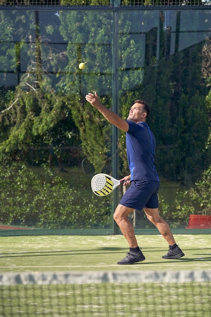 Vista lateral de cuerpo completo de un hombre activo con ropa deportiva lanzando pelota mientras juega pádel en el patio de juegos con red durante el entrenamiento