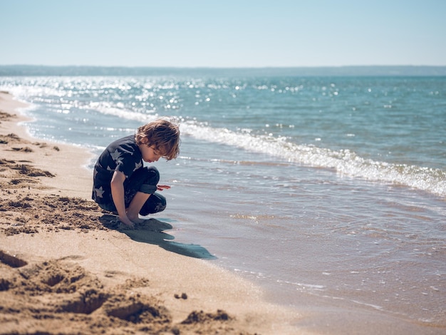 Vista lateral del cuerpo completo de un niño encantador sentado en el suelo y jugando con arena mojada cerca del mar ondulante en un día soleado de verano