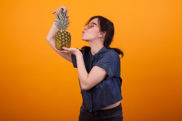Vista lateral da bela jovem beijando suas deliciosas ananás no estúdio sobre fundo amarelo. saborosa fruta exótica. mulher jovem e atraente com abacaxi.