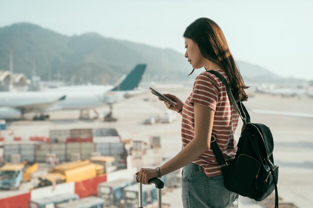 vista lateral da bela senhora de viagens com mochila e mala de bagagem caminhando para a sala de embarque no corredor. mulher turista em pé perto da janela usando o celular conversando aviões online na pista.