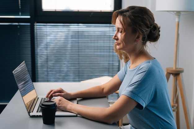 Vista lateral da empresária loira séria trabalhando no computador portátil olhando para a tela com expressão concentrada séria sentado na mesa