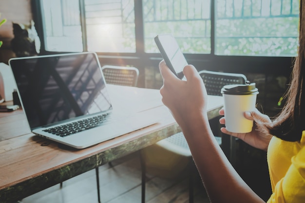 Vista lateral da encantadora jovem hipster usando o telefone inteligente e mãos segurando copo de café quente sentado na mesa de madeira em uma cafeteria. mulher usando laptop em um café - estilo de cor vintage.