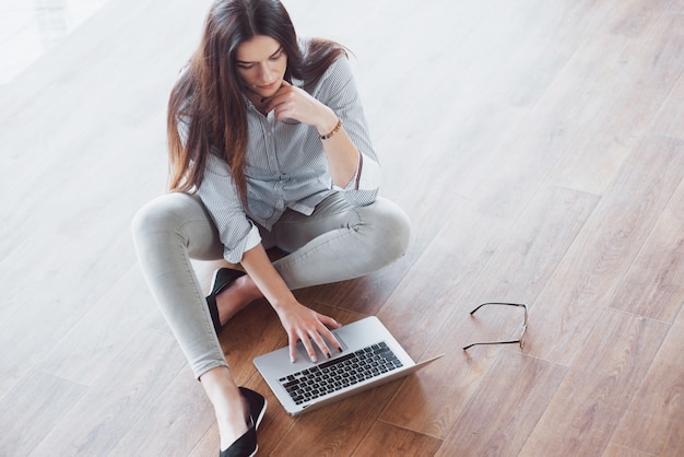 Vista lateral da garota atraente usando um laptop na área de wifi público e sorrindo enquanto está sentado no chão
