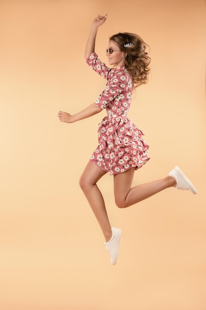 Vista lateral da garota louca usando vestido floral rosa e tênis branco pulando e rindo no estúdio Mulher jovem e atraente em óculos, aproveitando o clima quente e roupa elegante Conceito de felicidade