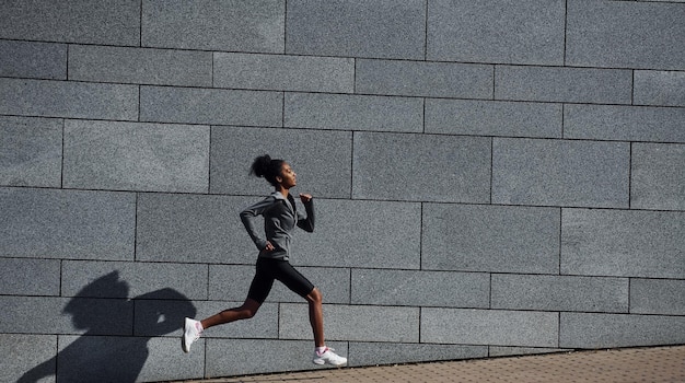 Vista lateral da jovem afro-americana em roupas esportivas que corre perto da parede