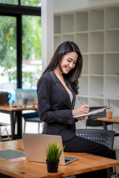 Vista lateral da jovem empresária asiática segurando a caneta uma anotação sentada à beira de uma mesa de laptop no escritório.