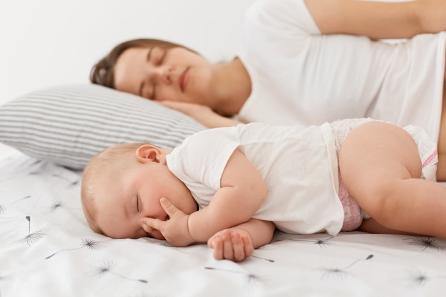 Vista lateral da mãe adulta jovem e seu bebê dormindo juntos, deitado na cama, posando em casa, na sala iluminada, a mamãe pode descansar enquanto a filha dorme.