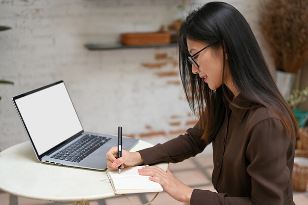 Vista lateral da mão feminina freelancer escrevendo no caderno enquanto trabalhava com o laptop na mesa redonda no café