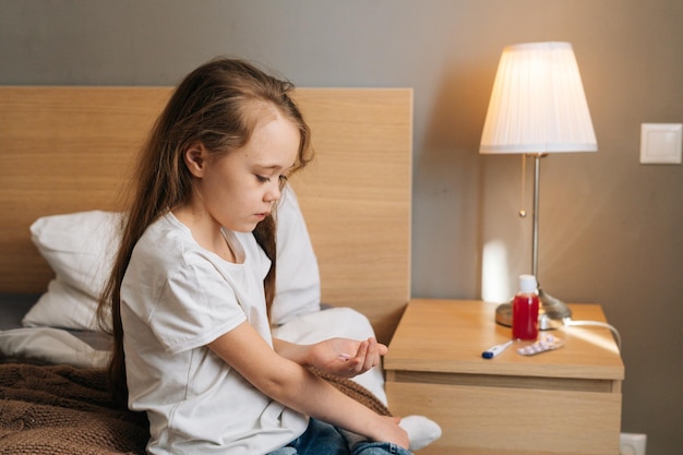 Vista lateral da menina doente segurando pílulas médicas nas mãos. Criança sentada na beira da cama com remédios em casa. Conceito de saúde infantil.