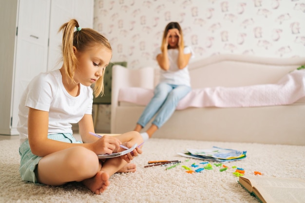 Vista lateral da menina primária escrevendo lição de casa no caderno sentado no chão com o laptop para educação a distância on-line em casa, no fundo desfocado da jovem mãe estressada sentada no sofá.