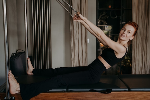 Foto vista lateral da mulher jovem ruiva bonita fazendo exercícios de alongamento do braço no reformador de pilates no estúdio de fitness, sorrindo no processo para aumentar a força dos músculos. estilo de vida saudável e fitness