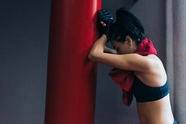 Vista lateral da mulher morena atraente desportiva com toalha vermelha no pescoço após o treino colocou a cabeça no saco de pancadas vermelho usando luvas de kickboxing na academia Esporte fitness lifestyle pessoas motivação