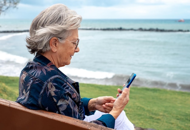 Vista lateral da mulher sênior sentada em um banco na praia em um dia de inverno usando telefone celular