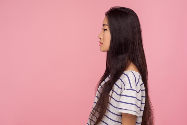 Vista lateral da séria calma linda garota asiática com longos cabelos morenos em camiseta listrada, olhando para o espaço da cópia com expressão pensativa concentrada. tiro de estúdio interior isolado no fundo rosa