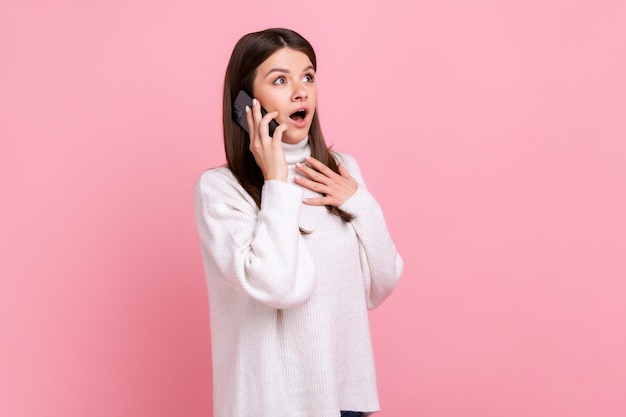 Vista lateral da surpreendida mulher falando ao telefone ouvindo notícias de última hora mantendo a boca aberta vestindo suéter branco estilo casual Tiro de estúdio interno isolado no fundo rosa