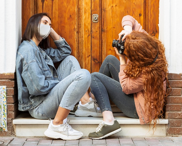 Foto vista lateral de amigas com máscaras sentadas ao lado da porta tirando fotos com a câmera