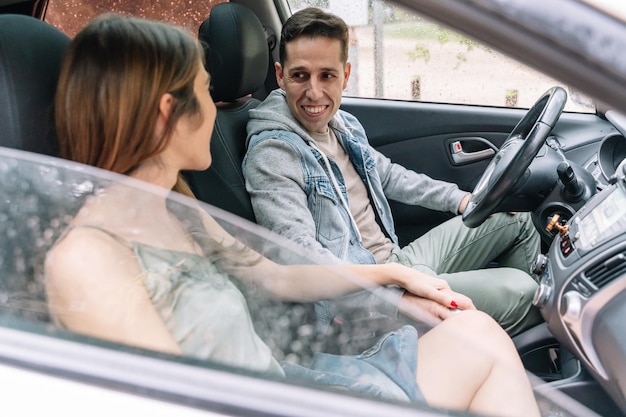 Vista lateral de casal alegre, sorrindo dentro de uma cadeirinha. Retrato horizontal de homem caucasiano viajando de carro em um dia chuvoso. Conceito de pessoas e viagens.