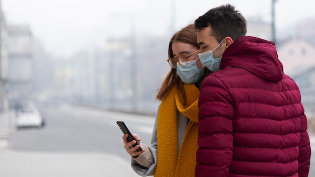 Foto vista lateral de casal usando smartphone na cidade, usando máscara médica