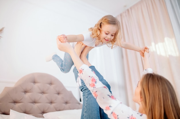 Vista lateral de comprimento total mãe jovem feliz, deitado na cama, levantando a filha criança pré-escolar. garota garoto pequeno fazendo avião, se divertindo com mamãe forte juntos no quarto