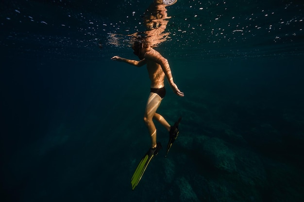 Vista lateral de corpo inteiro de mergulhador masculino sem camisa irreconhecível em nadadeiras e óculos flutuando sob a água azul do mar profundo em dia ensolarado