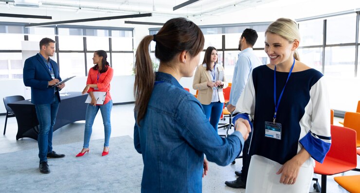 Foto vista lateral de duas empresárias caucasianas trabalhando em um escritório moderno e brilhante, em pé, conversando, sorrindo e apertando as mãos, com colegas de trabalho conversando em segundo plano