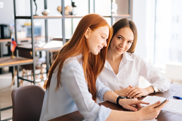 Vista lateral de duas mulheres de negócios jovens criativas trabalhando usando tablet digital na mesa de reunião com documentos de trabalho no escritório colegas de negócios usando computador touchscreen para discussão do projeto