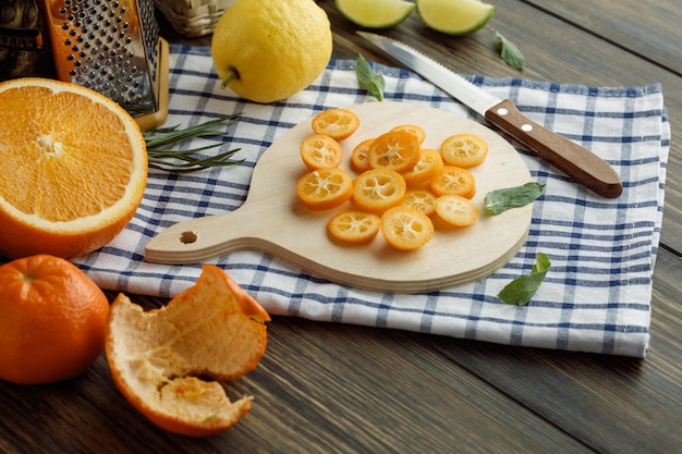 Vista lateral de fatias de kumquat na tábua com limão cortado faca ralador de cebolinha laranja em pano xadrez com fatias de limão tangerina com casca em fundo de madeira