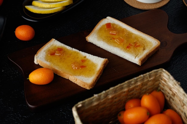 Vista lateral de fatias de pão com geléia manchada sobre eles na tábua com kumquats de fatias de limão na cesta em fundo preto