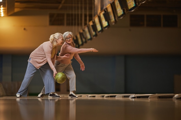 Vista lateral de grande angular para duas mulheres sêniors jogando boliche juntas enquanto desfrutam de entretenimento ativo na pista de boliche, copie o espaço