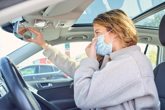 Vista lateral de mulher de casaco vestindo máscara médica enquanto toca o espelho retrovisor e olha para a frente no automóvel