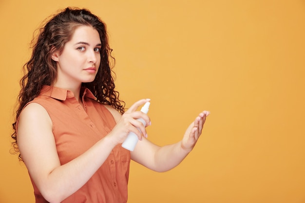 Vista lateral de mulher jovem com cabelo encaracolado usando anti-séptico para limpar as mãos em laranja, proteção durante a pandemia de coronavírus