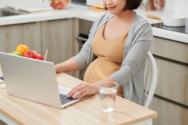 Vista lateral de perfil atraente e progressiva jovem mãe sentada na cadeira atrás da mesa dentro de um apartamento bem iluminado e usando o laptop