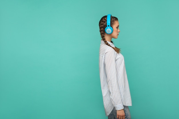 Vista lateral de perfil de menina em camisa azul listrada e penteado pigtail, ouvindo música em pé com fone de ouvido e desviando o olhar com uma cara séria, estúdio interno, isolado em fundo verde