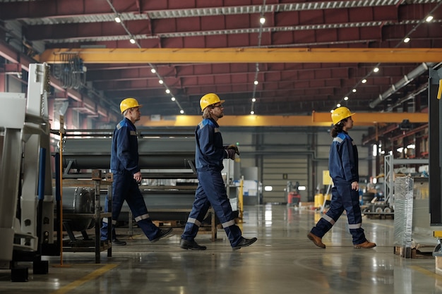 Vista lateral de três engenheiros de uma fábrica moderna ou planta industrial avançando enquanto vão para outra oficina ou para almoçar