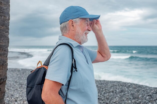 Vista lateral de um atraente homem barbudo sênior parado na praia olhando para as ondas e o horizonte sobre o mar Sorrindo um idoso relaxado aproveitando a aposentadoria ou as férias