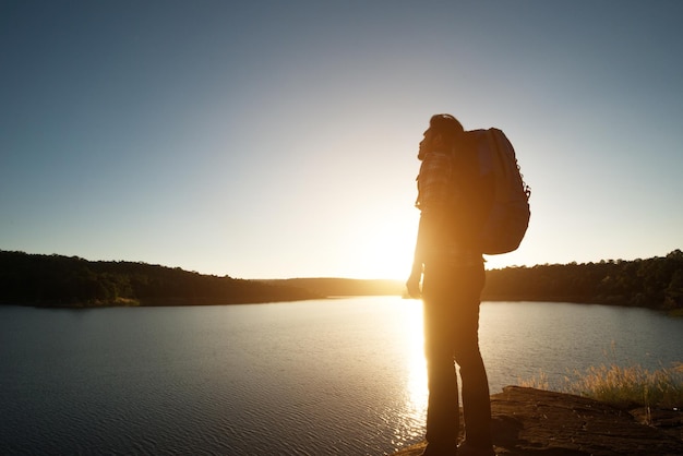 Foto vista lateral de um caminhante masculino carregando uma mochila enquanto está de pé na rocha à beira do lago contra um céu claro