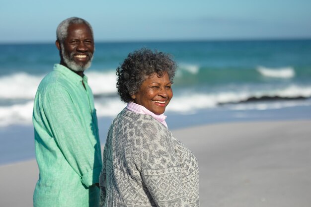 Vista lateral de um casal afro-americano idoso caminhando na praia com o céu azul e o mar ao fundo, de mãos dadas, virando-se e sorrindo para a câmera