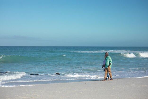 Vista lateral de um casal afro-americano sênior caminhando na praia com céu azul e mar ao fundo, de mãos dadas e conversando