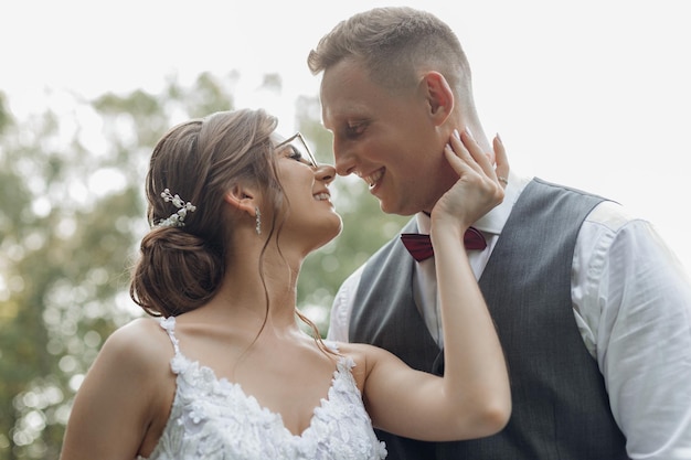 Vista lateral de um casal de noivos feliz e sorridente parado perto de árvores Jovem mulher bonita tocando o pescoço de um jovem